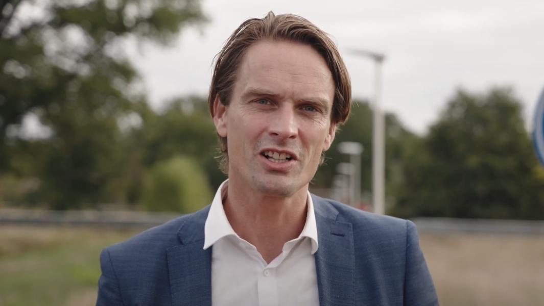 Man in blauw pak met wit overhemd buiten op een weg met groen op de achtergrond.