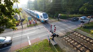 Trein nadert spoorwegovergang met wachtende auto's en gesloten slagbomen, omgeven door groen.