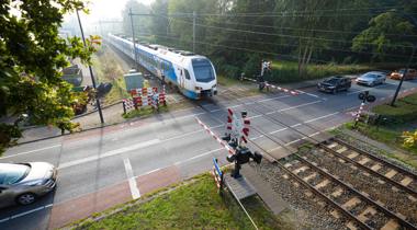 Trein nadert spoorwegovergang met wachtende auto's en gesloten slagbomen, omgeven door groen.