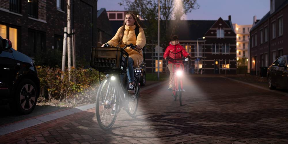 Twee fietsers, een jonge vrouw en een kind, fietsen in het donker door een woonwijk met hun fietsverlichting aan.