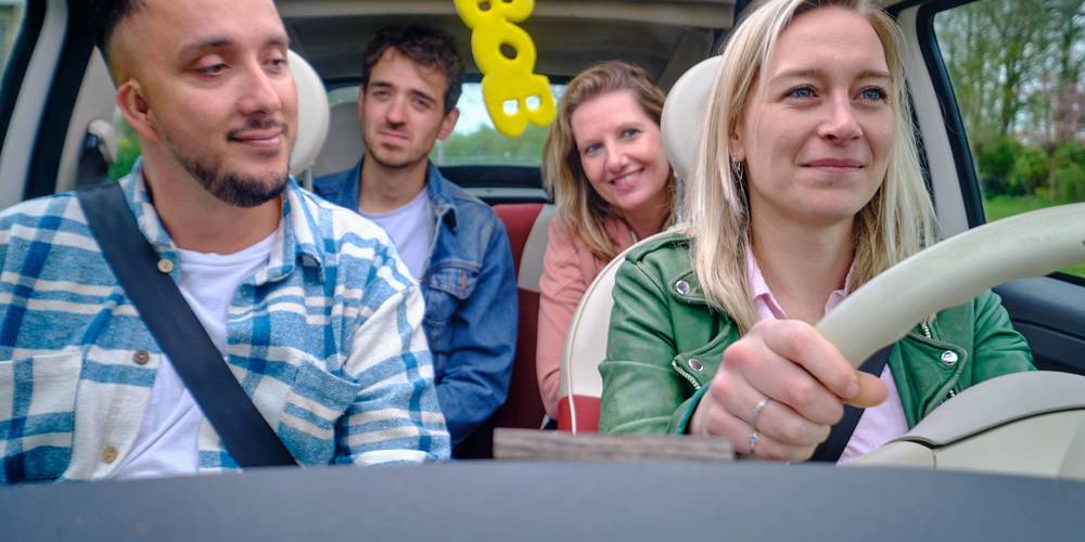 Vier vrolijke vrienden in een auto, met een gele BOB-sleutelhanger aan de binnenspiegel, kijken glimlachend naar voren terwijl een vrouw het stuur vasthoudt.