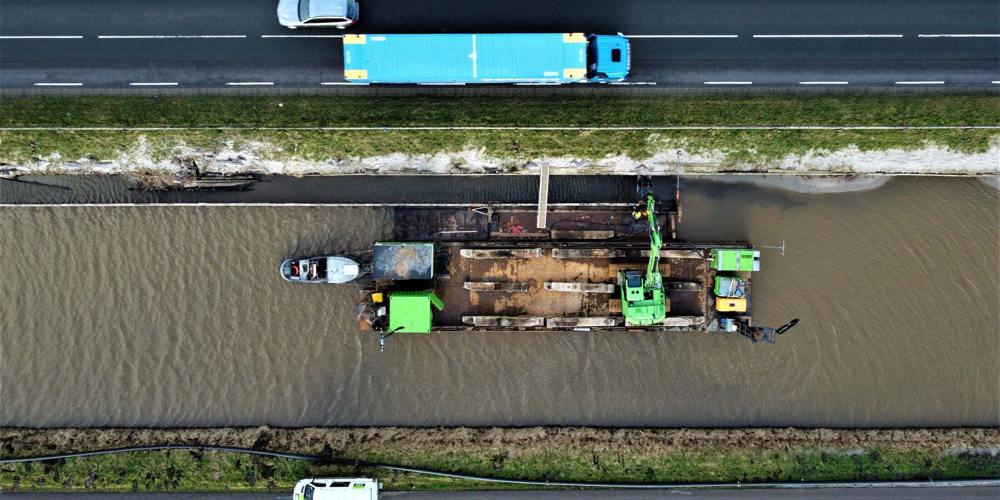  Een top-down luchtfoto van een vrachtwagen en een auto op een snelweg parallel aan een kanaal met een baggerschip en twee kleinere boten.