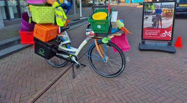 Overvolle fiets beladen met kleurrijke plastic spullen staat op straat naast een bord met de tekst: "Kort ritje naar de weekmarkt? Pak de fiets!" – promotie voor fietsen naar de markt.