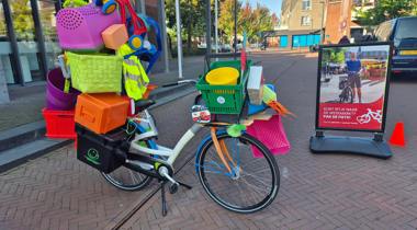 Overvolle fiets beladen met kleurrijke plastic spullen staat op straat naast een bord met de tekst: "Kort ritje naar de weekmarkt? Pak de fiets!" – promotie voor fietsen naar de markt.