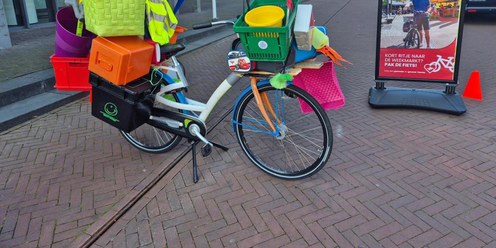 Overvolle fiets beladen met kleurrijke plastic spullen staat op straat naast een bord met de tekst: "Kort ritje naar de weekmarkt? Pak de fiets!" – promotie voor fietsen naar de markt.