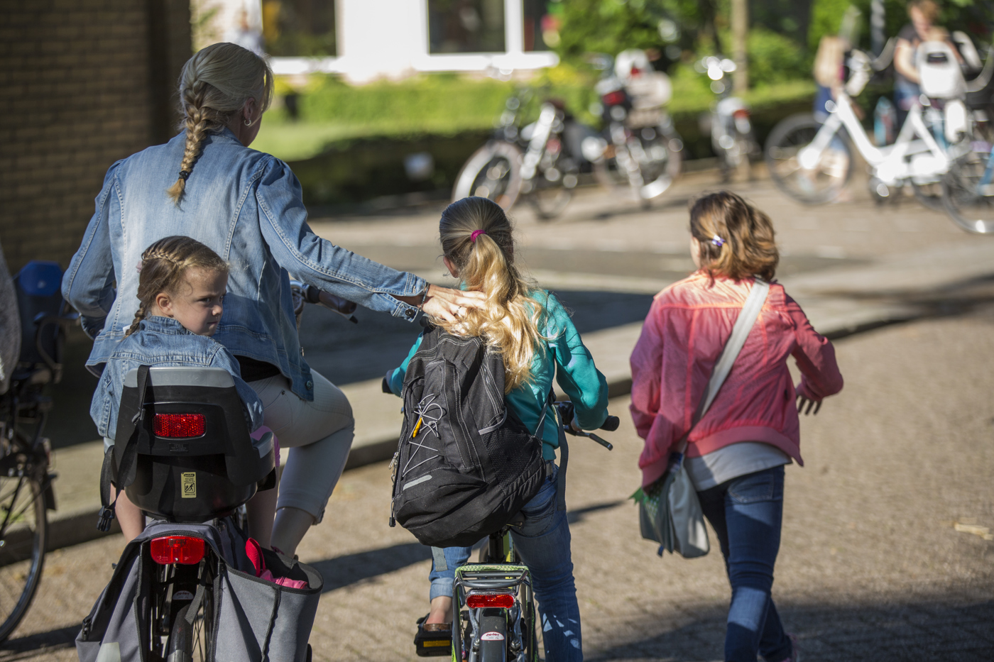 Een moeder op de fiets met een kind achterop en rechts naast haar rijdt ook een kind. Rechts van hen loopt ook een kind richting de school op de achtergrond. Daar staan meer fietsen.