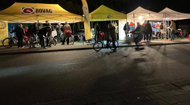 Mensen verzamelen zich bij verlichte tenten van BOVAG en ANWB tijdens een fietsverlichtingsactie in de avond.