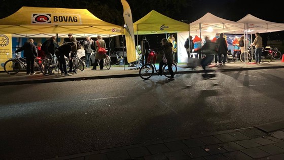 Mensen verzamelen zich bij verlichte tenten van BOVAG en ANWB tijdens een fietsverlichtingsactie in de avond.