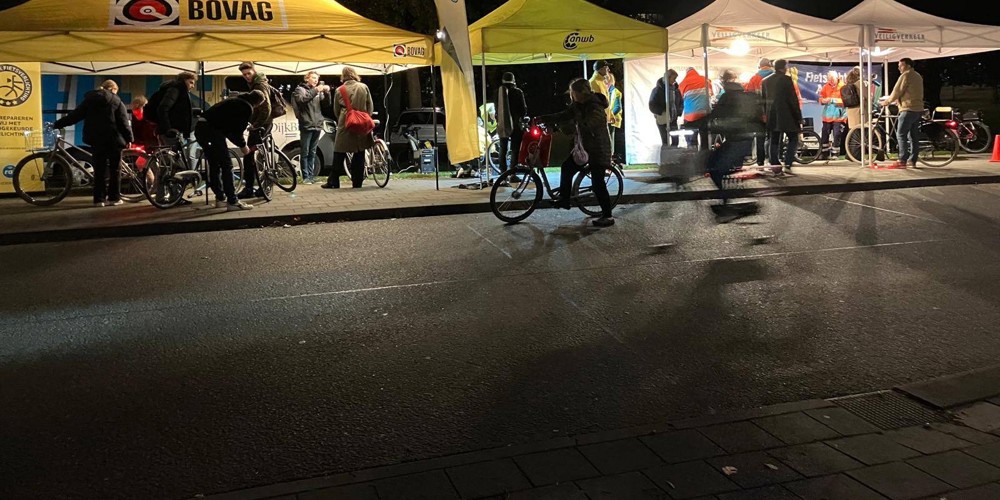 Mensen verzamelen zich bij verlichte tenten van BOVAG en ANWB tijdens een fietsverlichtingsactie in de avond.
