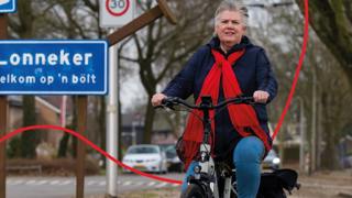 Een fietser, gekleed in een blauwe jas en een rode sjaal, rijdt op een fiets voor een blauw plaatsnaambord met "Lonneker" erop.