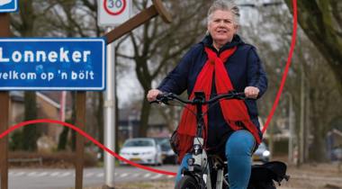 Een fietser, gekleed in een blauwe jas en een rode sjaal, rijdt op een fiets voor een blauw plaatsnaambord met "Lonneker" erop.