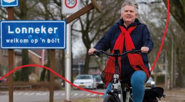 Een fietser, gekleed in een blauwe jas en een rode sjaal, rijdt op een fiets voor een blauw plaatsnaambord met "Lonneker" erop.