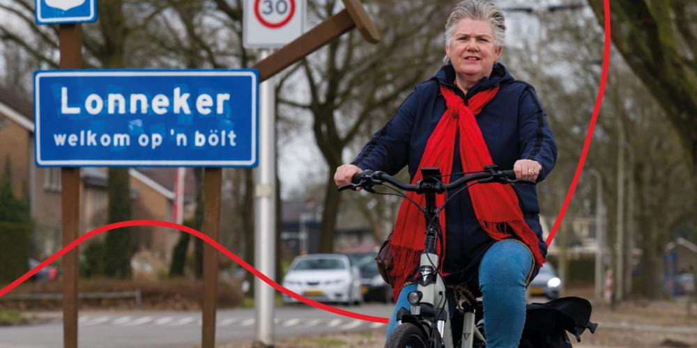 Een fietser, gekleed in een blauwe jas en een rode sjaal, rijdt op een fiets voor een blauw plaatsnaambord met "Lonneker" erop.