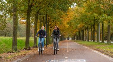  Twee meisjes fietsen naast elkaar op een fietspad omzoomd met bomen waarvan de bladeren beginnen te verkleuren in de herfst.