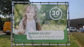 Een groot spandoek met een afbeelding van een lachend kind met een duim omhoog. Rechtsboven staat in een cirkel "30 KM/U". Onder de afbeelding staat de tekst "Dankjewel autorijder! Door jou kan ik veilig naar school".