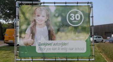Een groot spandoek met een afbeelding van een lachend kind met een duim omhoog. Rechtsboven staat in een cirkel "30 KM/U". Onder de afbeelding staat de tekst "Dankjewel autorijder! Door jou kan ik veilig naar school".