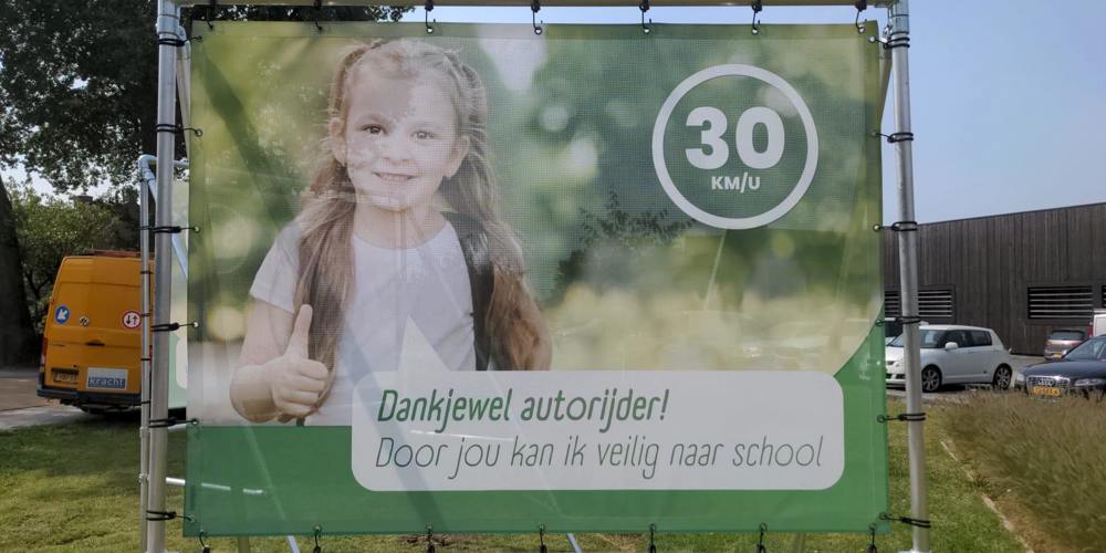 Een groot spandoek met een afbeelding van een lachend kind met een duim omhoog. Rechtsboven staat in een cirkel "30 KM/U". Onder de afbeelding staat de tekst "Dankjewel autorijder! Door jou kan ik veilig naar school".