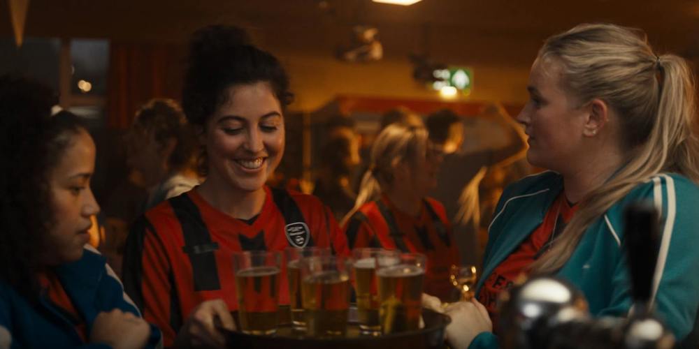 Drie vrouwen in sportkleding staan in een gezellige kantine; één van hen draagt een dienblad met glazen bier en glimlacht.