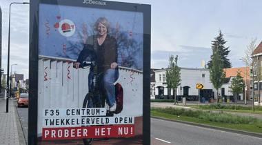 Een reclamebord langs de weg, met een afbeelding van een lachende vrouw op een elektrische fiets met de tekst "F35 CENTRUM - TWEKKELERVELD OPEN - PROBEER HET NU!
