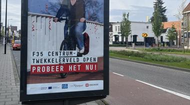 Een reclamebord langs de weg, met een afbeelding van een lachende vrouw op een elektrische fiets met de tekst "F35 CENTRUM - TWEKKELERVELD OPEN - PROBEER HET NU!