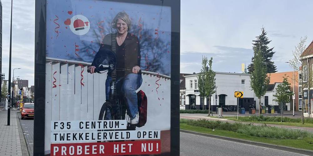 Een reclamebord langs de weg, met een afbeelding van een lachende vrouw op een elektrische fiets met de tekst "F35 CENTRUM - TWEKKELERVELD OPEN - PROBEER HET NU!