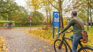 Twee fietsers rijden over een herfstachtig fietspad, omringd door bomen en met bladeren op de grond.