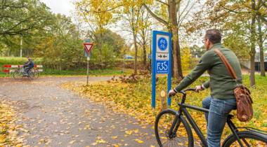 Twee fietsers rijden over een herfstachtig fietspad, omringd door bomen en met bladeren op de grond.