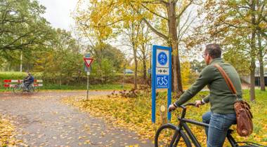 Twee fietsers rijden over een herfstachtig fietspad, omringd door bomen en met bladeren op de grond.