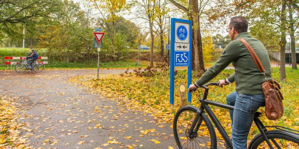 Twee fietsers rijden over een herfstachtig fietspad, omringd door bomen en met bladeren op de grond.