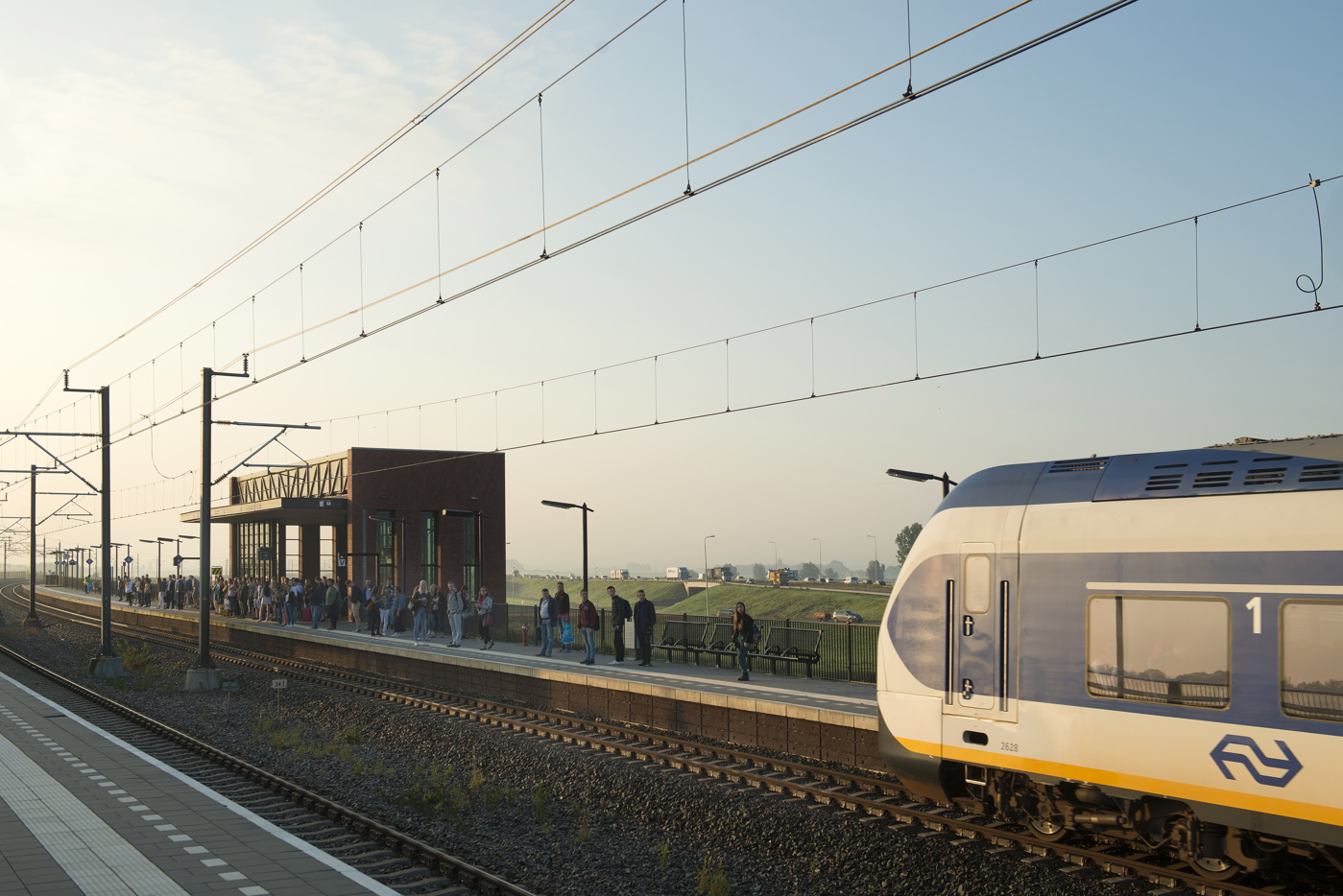 Een trein arriveert bij Station Kampen-Zuid. Op het perron staan mensen te wachten. Op de achtergrond rijden auto's op de provinciale weg N50.