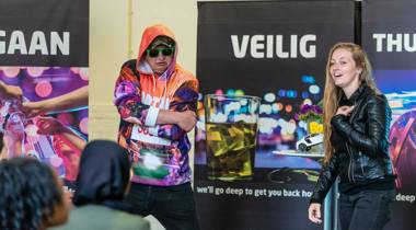 Een vrouw in een leren jack lacht terwijl een man in een kleurrijke hoodie en zonnebril poseert. Ze staan voor banners met de tekst "VEILIG THUIS".