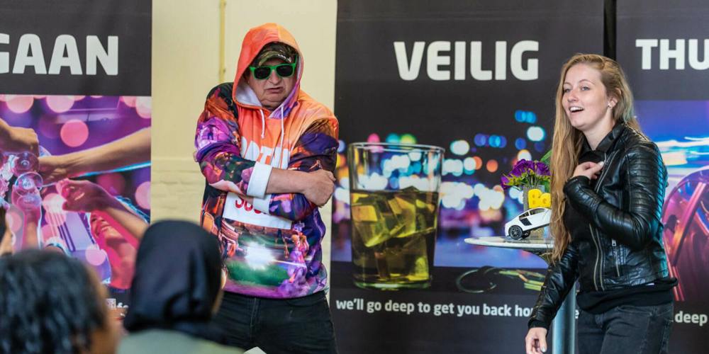 Een vrouw in een leren jack lacht terwijl een man in een kleurrijke hoodie en zonnebril poseert. Ze staan voor banners met de tekst "VEILIG THUIS".