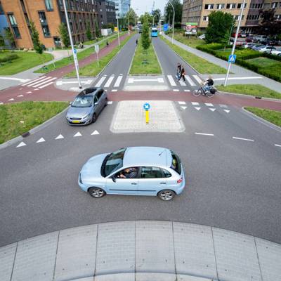 Een auto nadert een rotonde in een stedelijke omgeving, terwijl een fietser en voetgangers de weg oversteken.