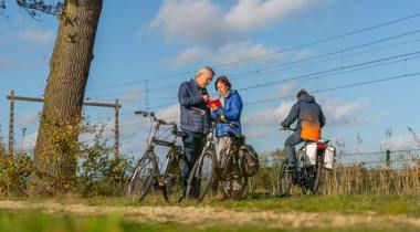 Twee oudere mensen op fietsen stoppen om naar een kaart te kijken, terwijl een derde fietser verderop op het pad rijdt.