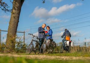 Twee oudere mensen op fietsen stoppen om naar een kaart te kijken, terwijl een derde fietser verderop op het pad rijdt.