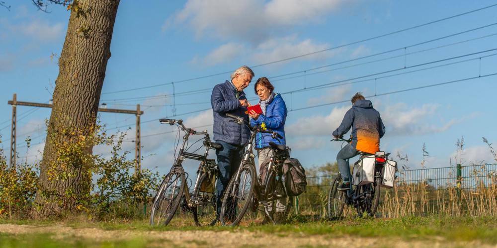 Twee oudere mensen op fietsen stoppen om naar een kaart te kijken, terwijl een derde fietser verderop op het pad rijdt.