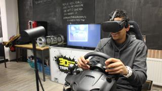 Persoon met VR-headset en stuurcontroller simuleert autorijden in kamer met monitor, speakers en apparatuur.