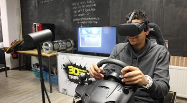 Persoon met VR-headset en stuurcontroller simuleert autorijden in kamer met monitor, speakers en apparatuur.