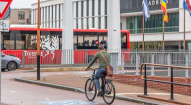 Fietser met groen vest en bruine tas op fietspad F35, met rode bus en stadhuis op de achtergrond.