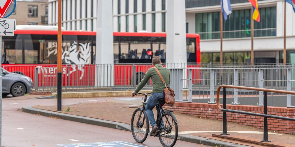 Fietser met groen vest en bruine tas op fietspad F35, met rode bus en stadhuis op de achtergrond.