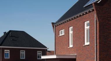 Een schuin omhoog gericht beeld van twee moderne bakstenen huizen met donkere daken, waarvan er één zonnepanelen heeft, onder een helderblauwe lucht.