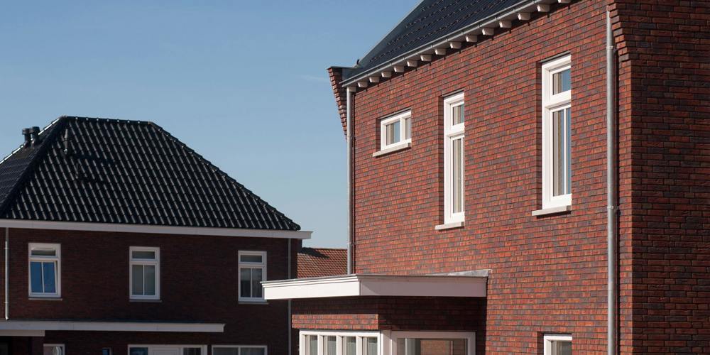 Een schuin omhoog gericht beeld van twee moderne bakstenen huizen met donkere daken, waarvan er één zonnepanelen heeft, onder een helderblauwe lucht.