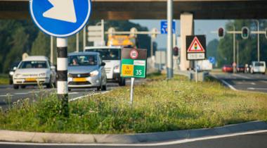 Drukke weg met verkeersborden, waaronder een blauw bord met witte pijl naar links, snelheidslimiet van 80 km/u, en een viaduct.