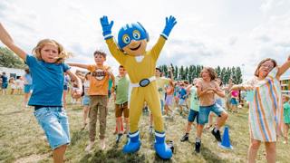 Kinderen springen blij in de lucht op een grasveld samen met een vrolijke mascotte in geel-blauw pak.