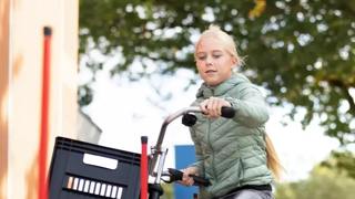 Een meisje op een fiets met een krat voorop, fietst tussen rode paaltjes door.