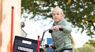 Een meisje op een fiets met een krat voorop, fietst tussen rode paaltjes door.