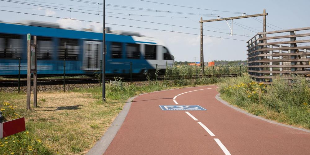 Blauwe trein rijdt langs een fietspad onder een blauwe lucht.