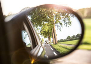 Een blik in een zijspiegel van een auto die een weg met bomen en een achteropkomende vrachtwagen laat zien, met een zonnige gloed.