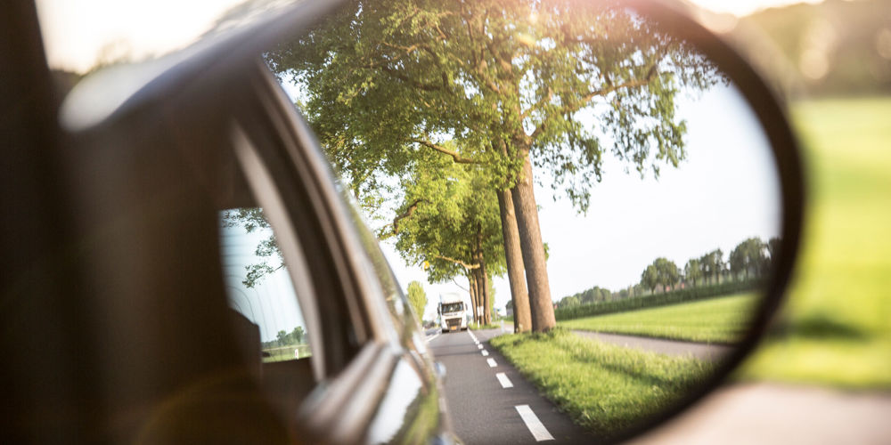 Een blik in een zijspiegel van een auto die een weg met bomen en een achteropkomende vrachtwagen laat zien, met een zonnige gloed.