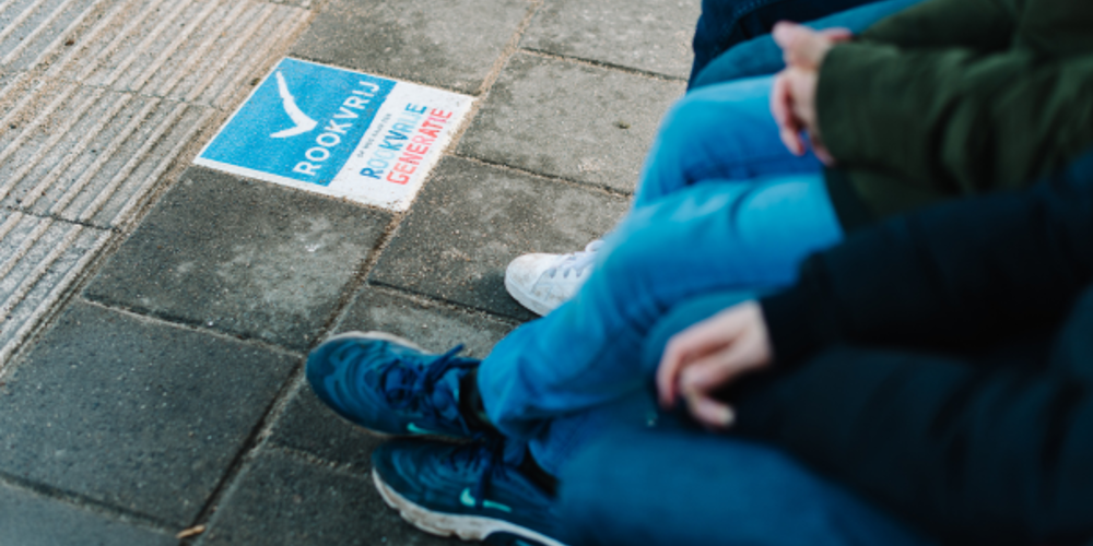 Groep mensen zittend bij rookvrije zone met tegeltekst 'ROOKVRIJ RUIMTE GEMEENTE'